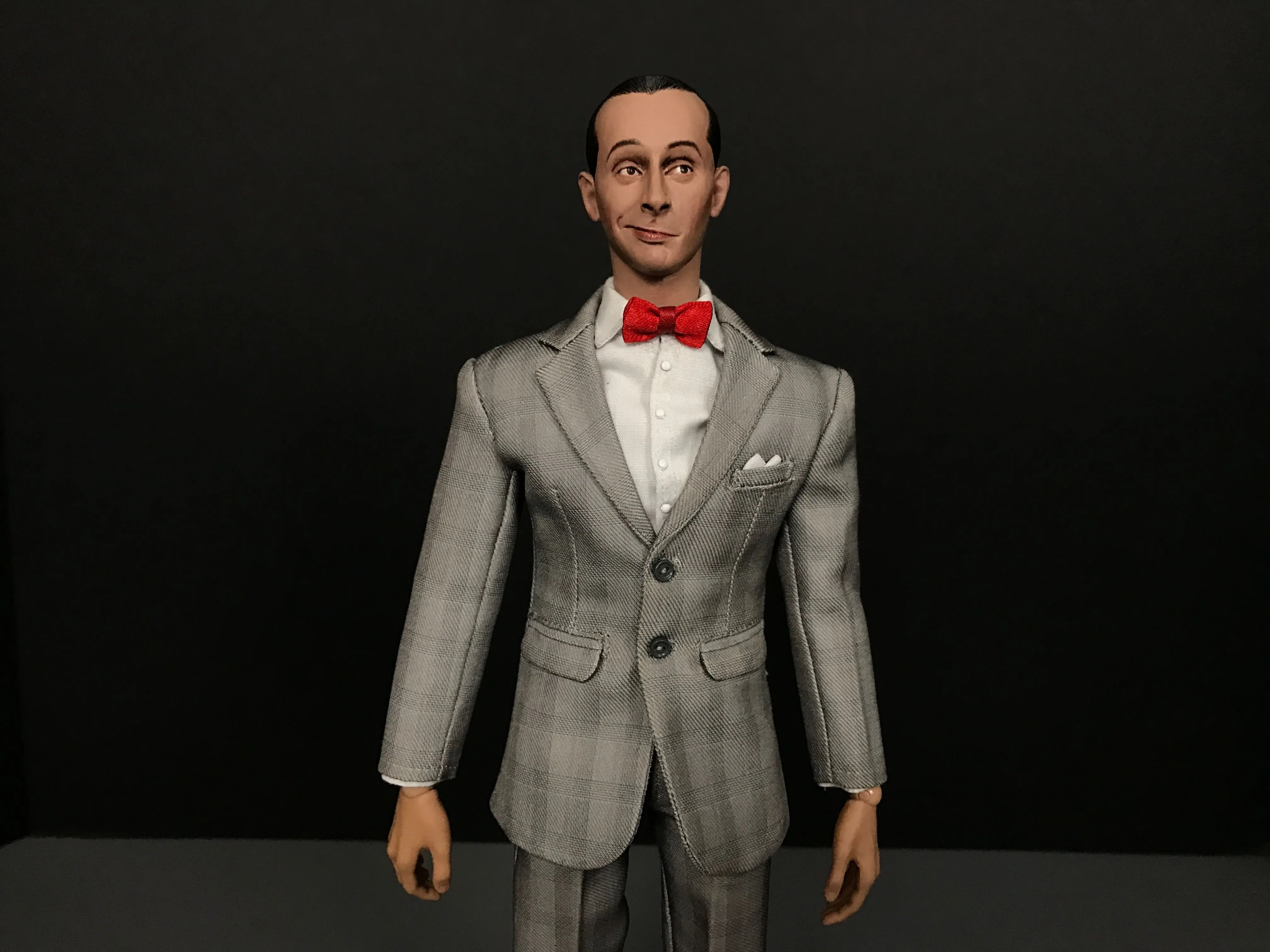 Custom Pee-wee Herman figure with a slight smile, gray plaid suit, white shirt and red bow tie against a dark background.