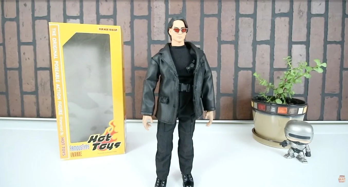 Hot Toys Famous Type Ethan Hunt standing outside the yellow box in a black jacket and trousers, with red-lensed sunglasses and a shoulder holster.