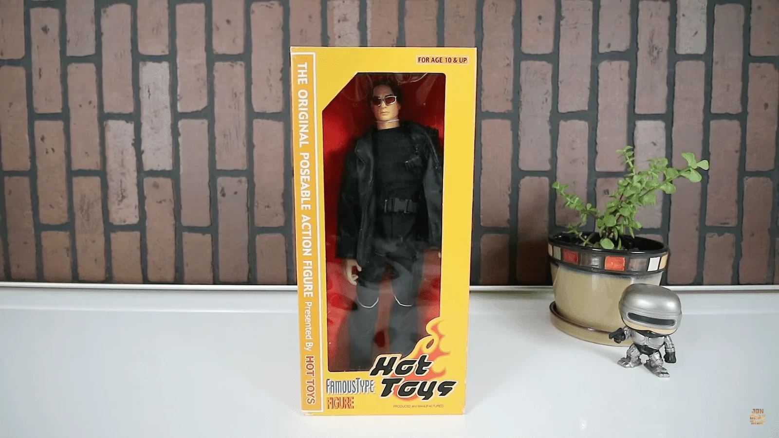 Early Hot Toys Famous Type Ethan Hunt figure in a yellow window box, wearing a black outfit and sunglasses beside a small plant and miniature RoboCop display.
