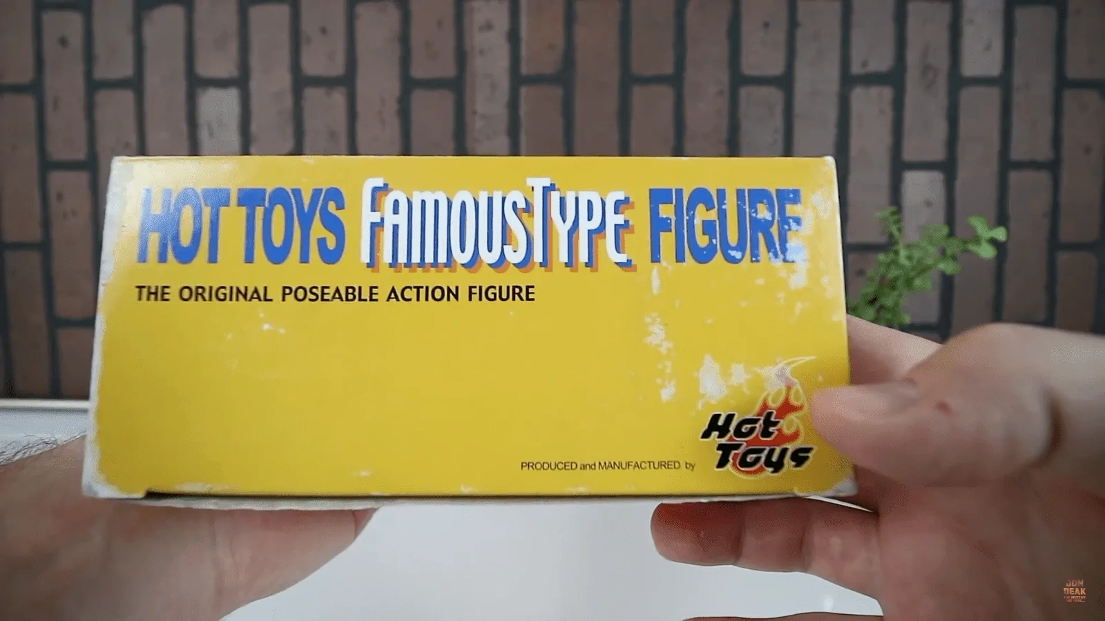 Hands holding the worn yellow box panel labelled Hot Toys Famous Type Figure and The Original Poseable Action Figure, with the Hot Toys flame logo.