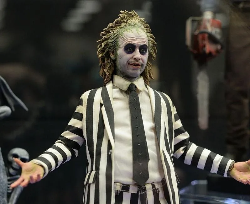 Beetlejuice figure with messy green-tinged hair, dark eye makeup and outstretched hands, wearing a black-and-white striped suit.