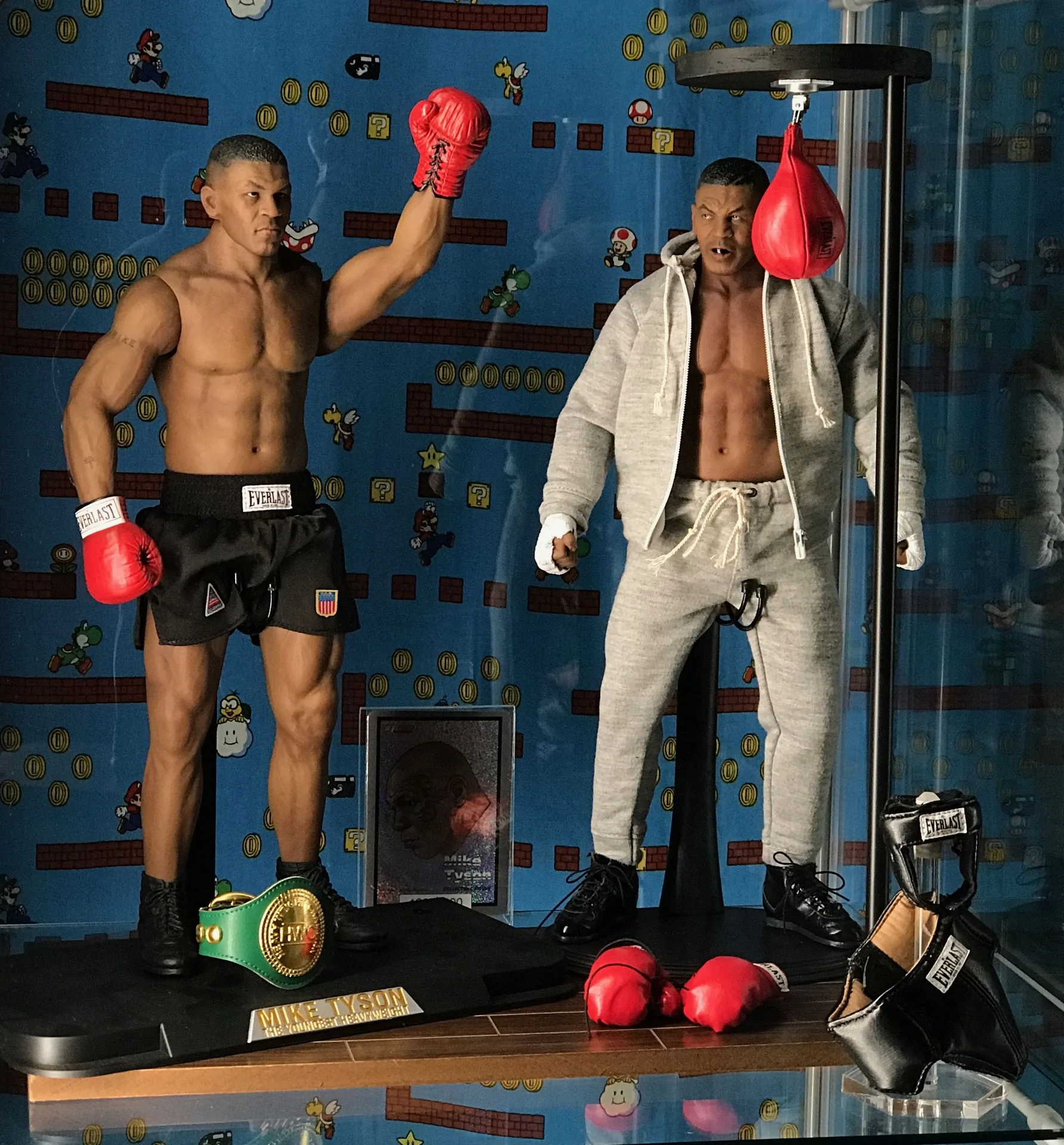 Two Storm Collectibles Mike Tyson figures displayed together: one in black boxing shorts and red gloves, the other in a grey tracksuit beside a speed bag, with a championship belt below.