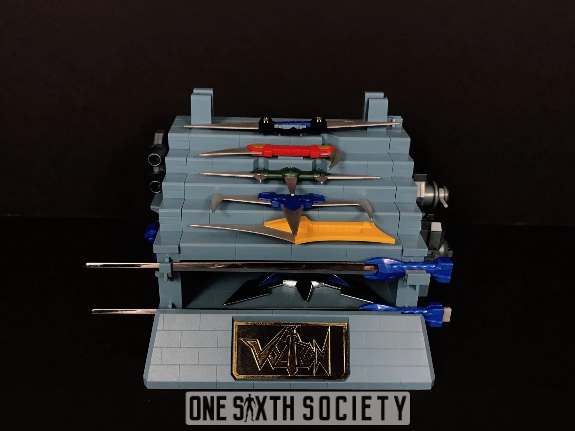 Gray stepped Voltron accessory stand holding color-coded lion weapons and longer swords above a black-and-gold Voltron nameplate.