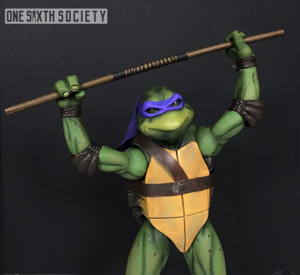 NECA Donatello holding his long bo staff above his head with both arms raised, wearing a purple mask and brown harness.