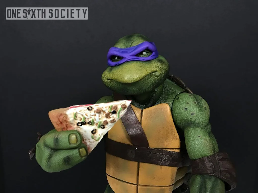 NECA Donatello holding a miniature pizza slice near his face while wearing his purple mask and brown chest harness.