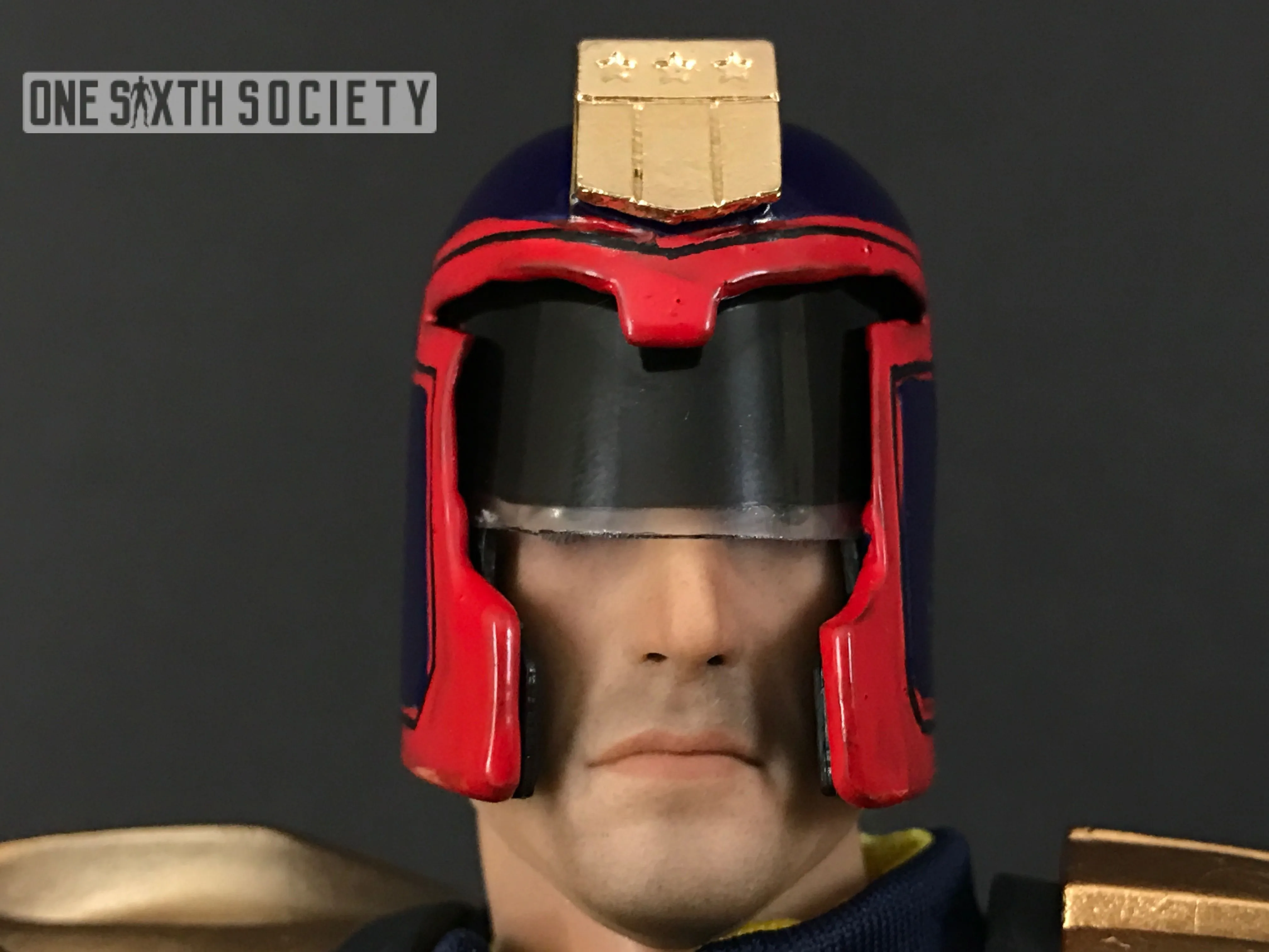 Close-up of the Judge Dredd figure's gold helmet badge, black visor, red trim and exposed lower face.