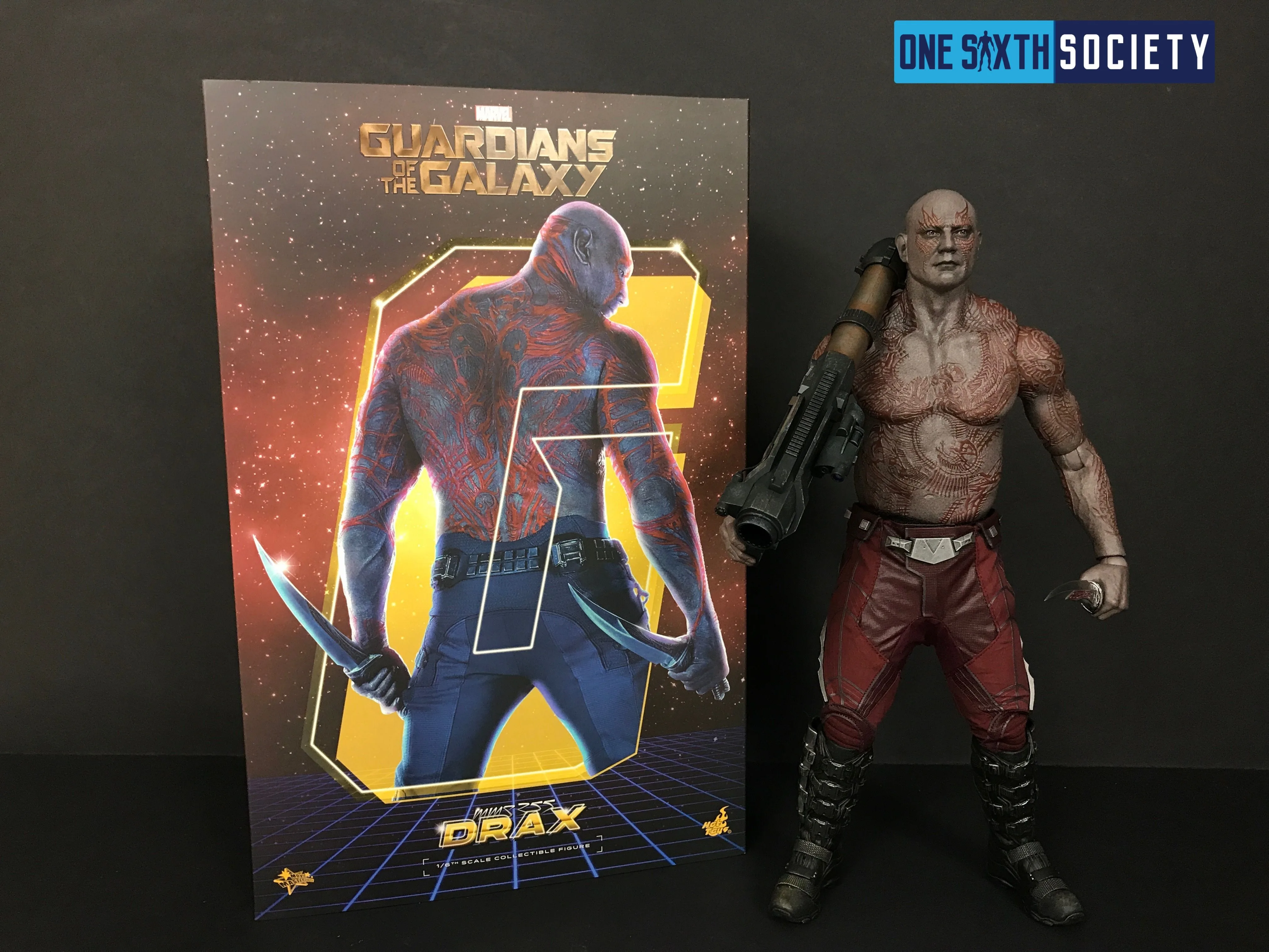 Drax in red trousers carrying a large weapon beside a neon-colored Guardians of the Galaxy Drax art box.