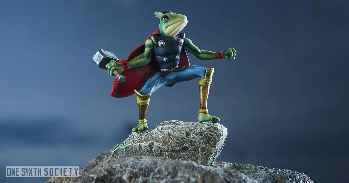 Green Frog Thor collectible holding a small hammer, wearing a red cape and gold-colored boots while posed on a rock.