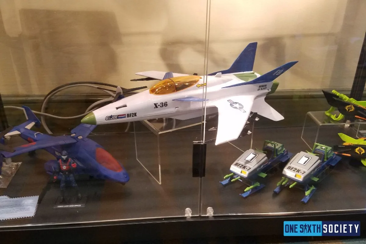 White X-36 toy aircraft with blue tail fins and an amber canopy displayed above smaller vehicles at JoeCon 2017.