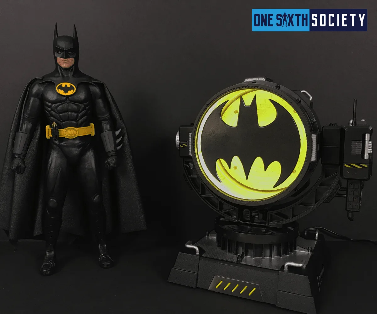 Batman figure in a black suit with a yellow belt and oval chest emblem beside an illuminated yellow Bat-Signal spotlight.