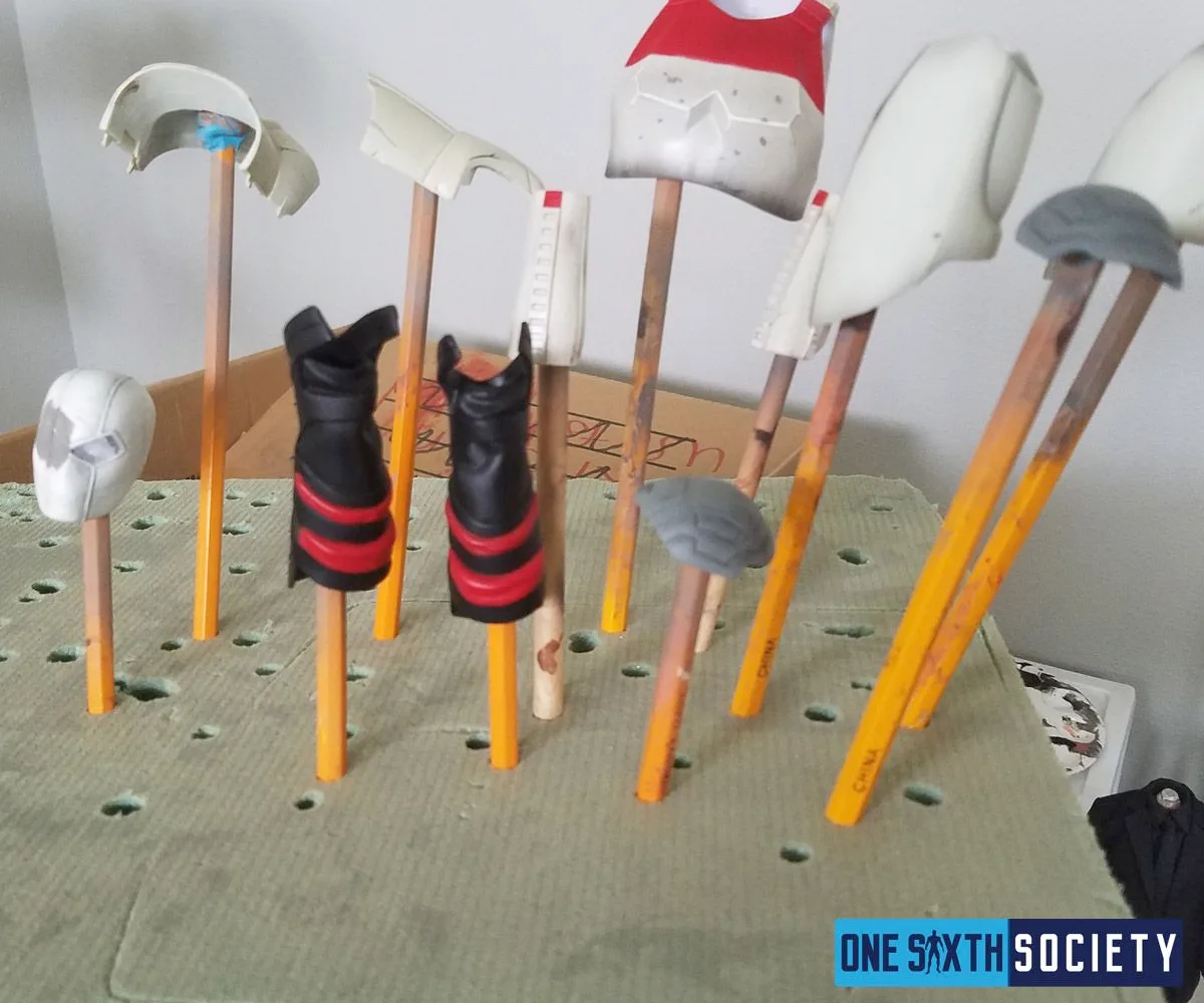Separate white, gray and red armor pieces and black-and-red boots mounted on wooden sticks in a green foam block.