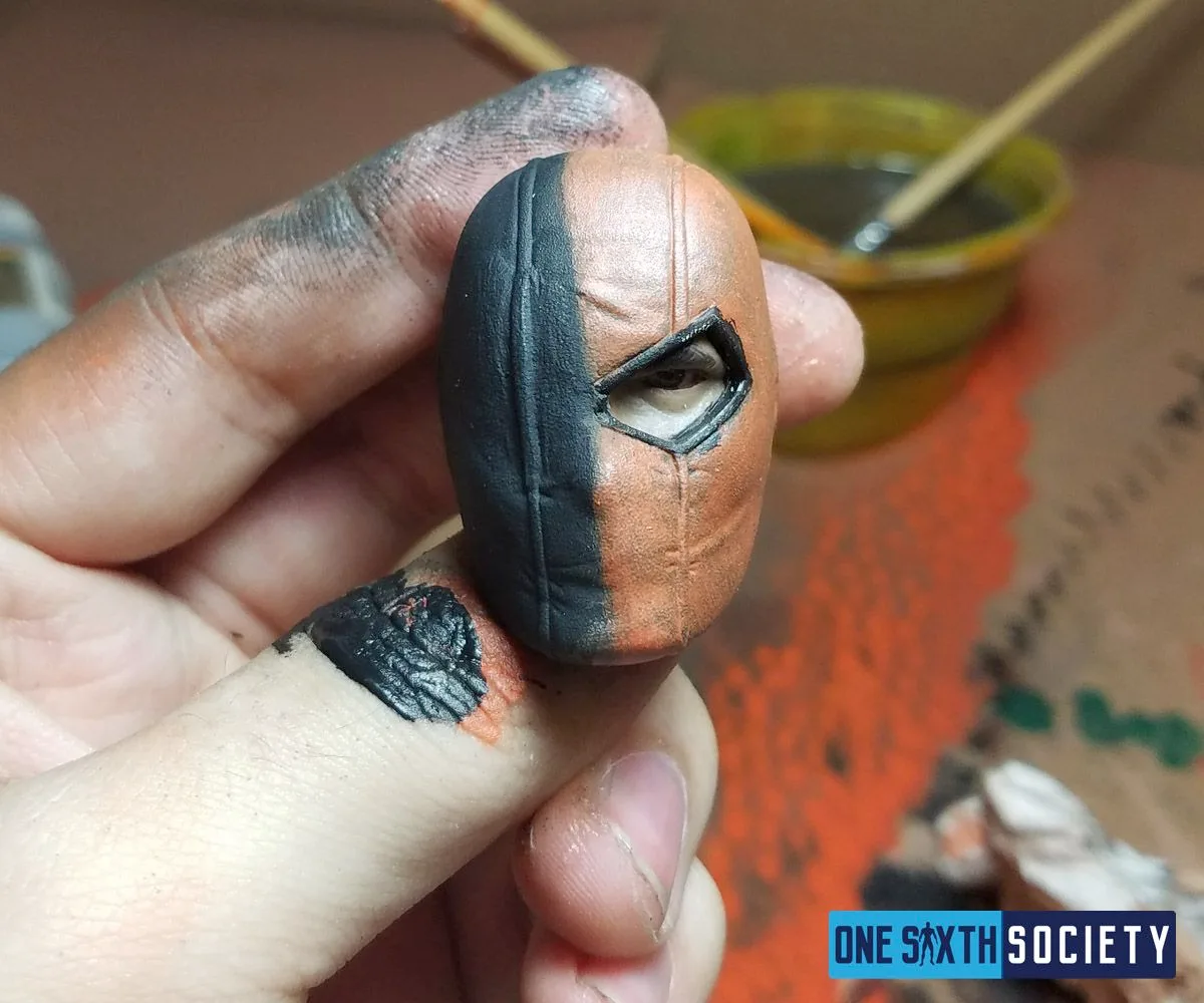 Paint-stained fingers holding the finished Deathstroke mask, divided into black and metallic-orange halves.