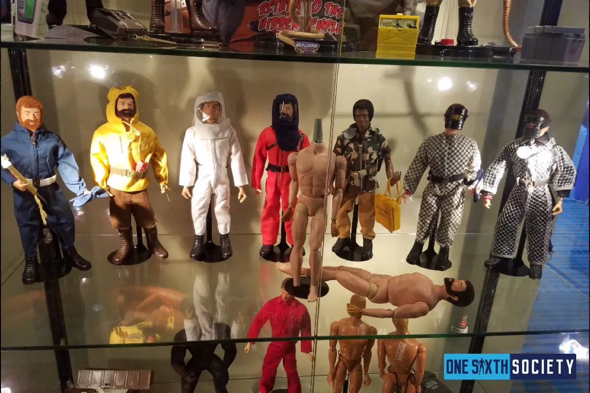G.I. Joe adventure figures in assorted uniforms stand behind two unclothed articulated figure bodies in a JoeCon 2017 display case.