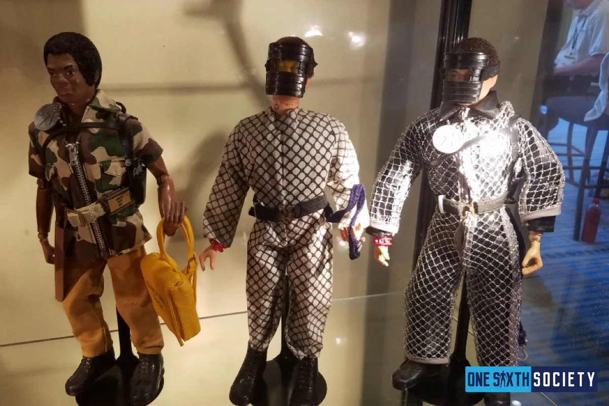 A camouflage-clad G.I. Joe figure stands beside two figures in patterned protective suits and dark face shields.
