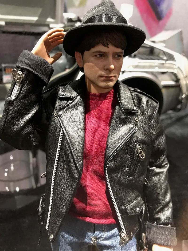 Waist-up view of Hot Toys Marty McFly raising a hand to his hat, wearing a textured black jacket, red sweater and blue jeans.