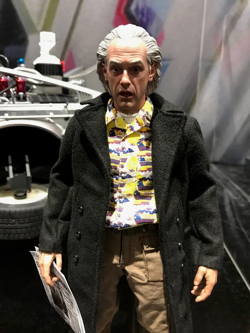 Hot Toys Doc Brown wearing a long dark coat over a colorful shirt and tan trousers, holding a newspaper at his side.