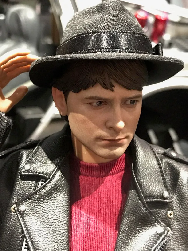 Close-up of Hot Toys Marty McFly beneath a dark brimmed hat, with one hand lifted toward it and a red shirt under his black jacket.