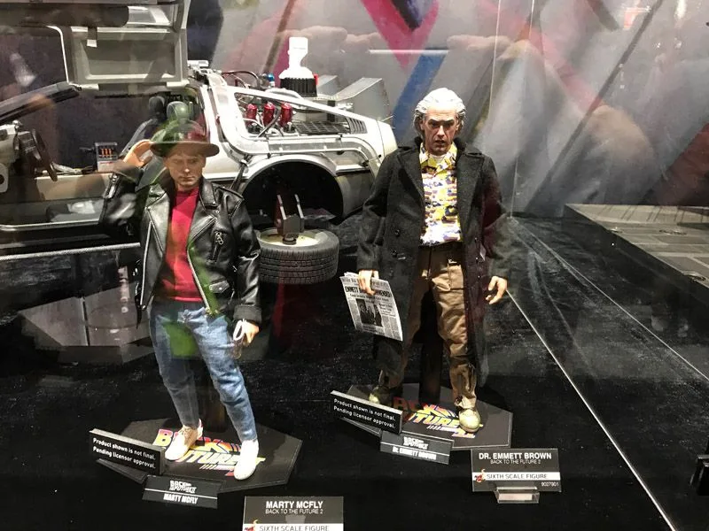 Hot Toys Marty McFly in a black jacket and hat beside Doc Brown in a dark coat, with the DeLorean time machine displayed behind them.