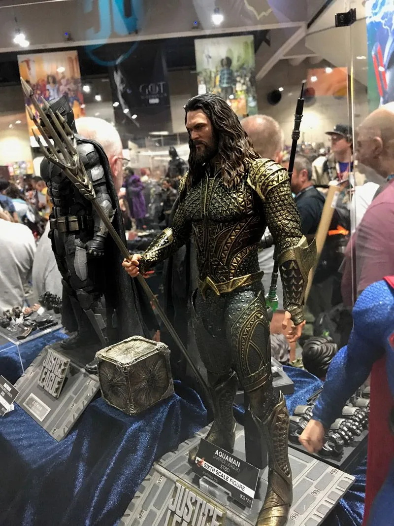 Hot Toys Aquaman standing on a rocky display base while holding a long trident, with his gold-and-green armor visible full length.