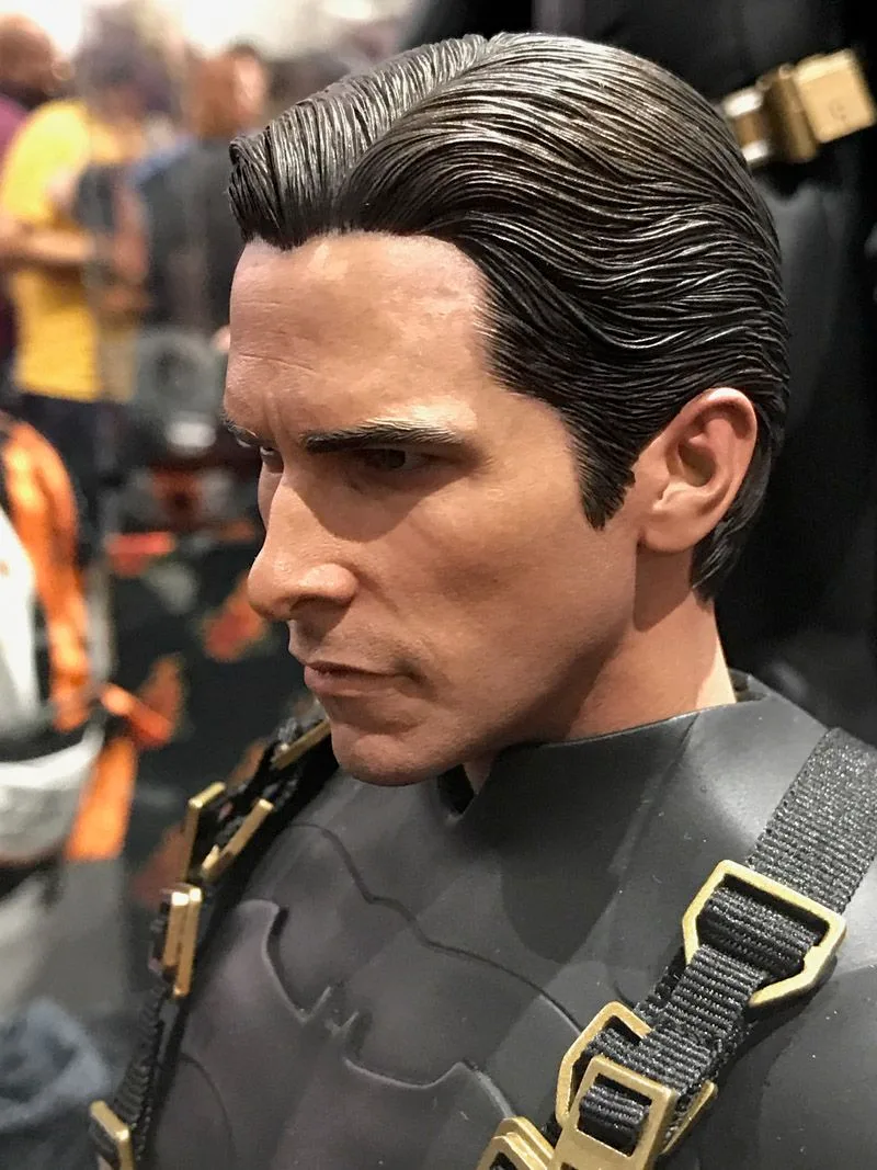 Close three-quarter profile of the Hot Toys Bruce Wayne head sculpt, with slicked-back dark hair and a gold-buckled shoulder strap.