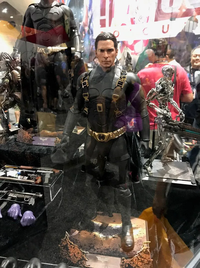 Hot Toys unmasked Bruce Wayne in a dark armored suit with gold-buckled shoulder straps, standing beside an accessory tray.