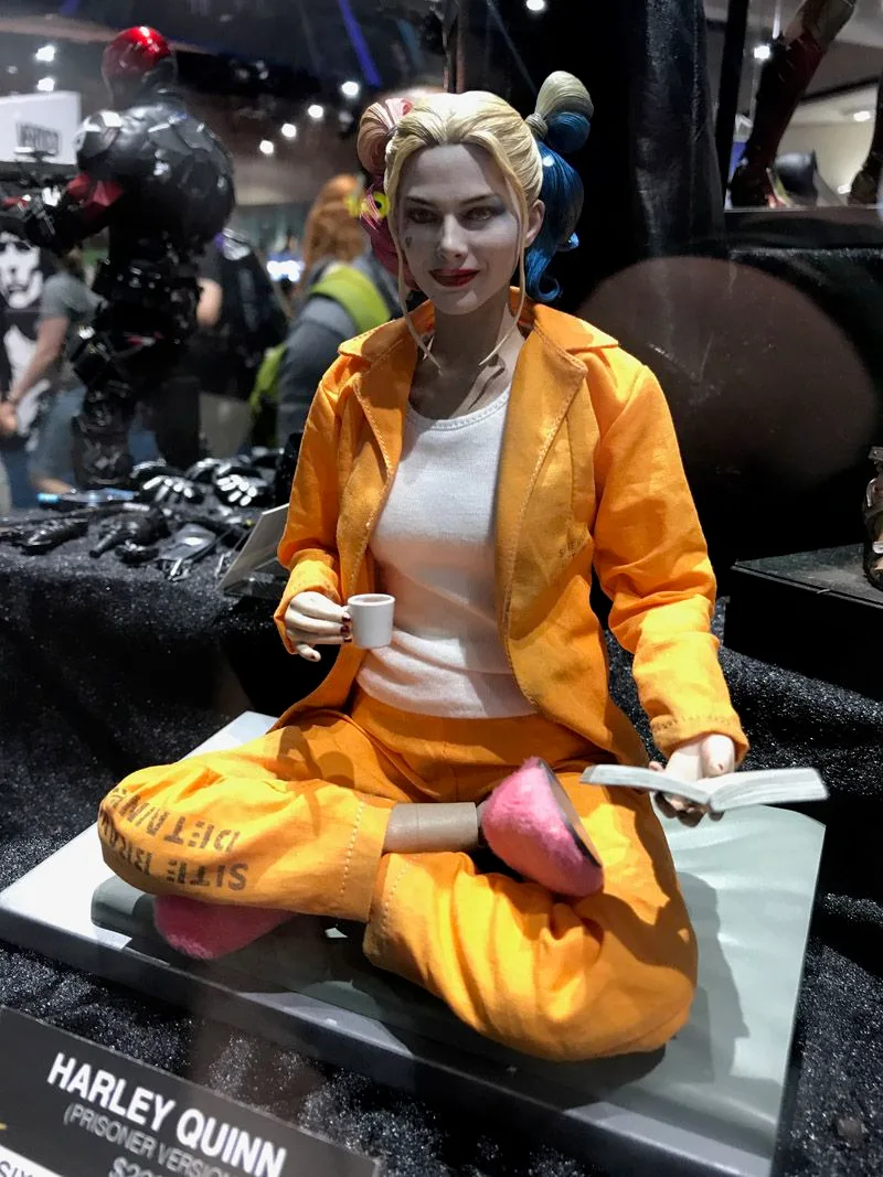 Hot Toys Harley Quinn sitting cross-legged in an orange prison outfit and pink slippers, holding a tiny cup beside an open book.