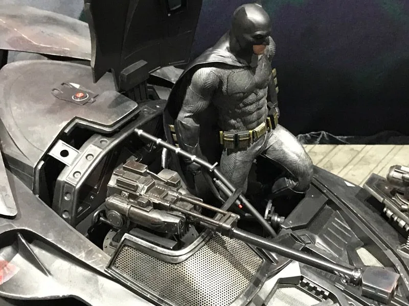 Side view of Hot Toys Batman kneeling at the Batmobile cockpit, with the long front gun assembly and exposed mechanical panels in the foreground.