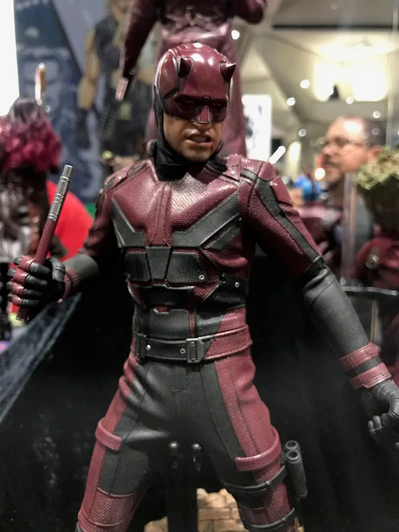 Hot Toys Daredevil wearing a dark red-and-black suit and horned mask, holding a baton upright.