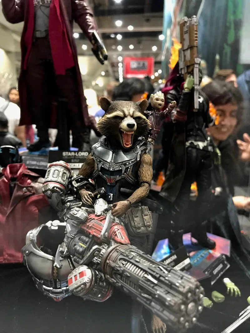 Hot Toys Rocket grinning behind an oversized multi-barreled weapon, with a small Groot perched behind his shoulder.