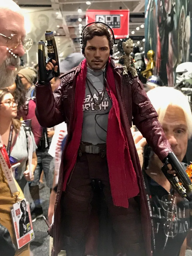 Hot Toys Star-Lord wearing headphones, a red scarf and long coat, holding two blasters with a small Groot on his shoulder.