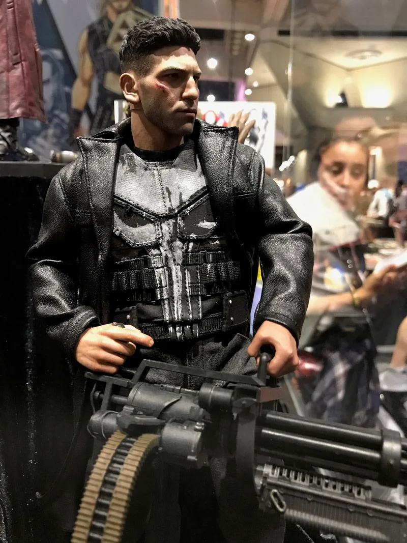 Hot Toys Punisher wearing a black coat and white skull-marked vest, holding a large multi-barreled weapon with an ammunition belt.