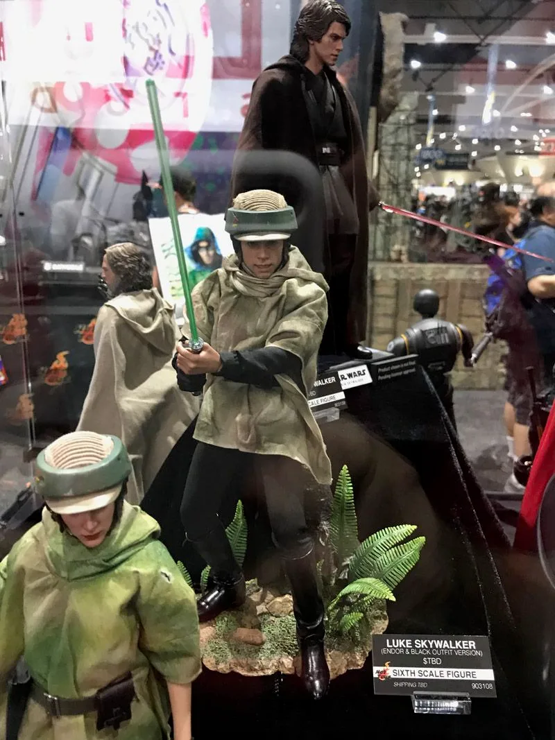 Hot Toys Luke Skywalker in an Endor helmet and camouflage poncho, holding a green lightsaber above a forest-themed base with ferns.