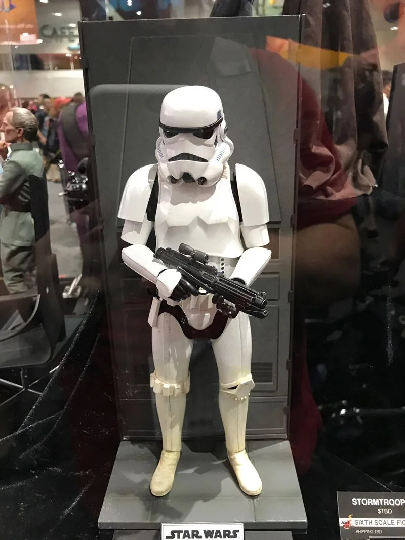 Hot Toys white-armored Stormtrooper holding a black blaster across his waist, shown full length against a gray display wall.