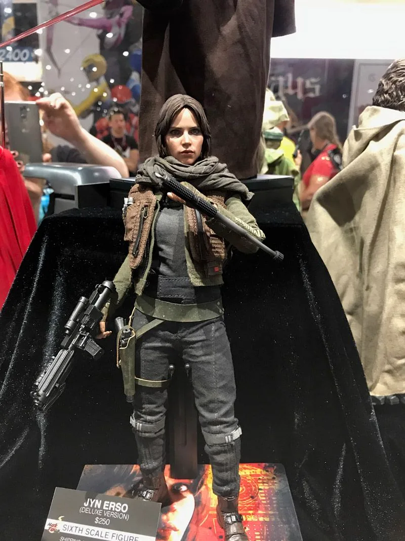 Hot Toys Jyn Erso in layered gray-green clothing, a brown vest and scarf, carrying a long blaster in the Rogue One display.