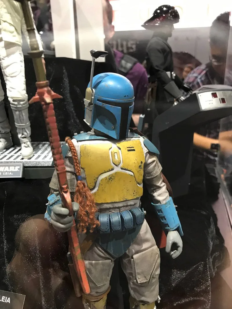 Hot Toys Boba Fett wearing a blue helmet, yellow chest plate and blue belt pouches, holding a long reddish weapon.