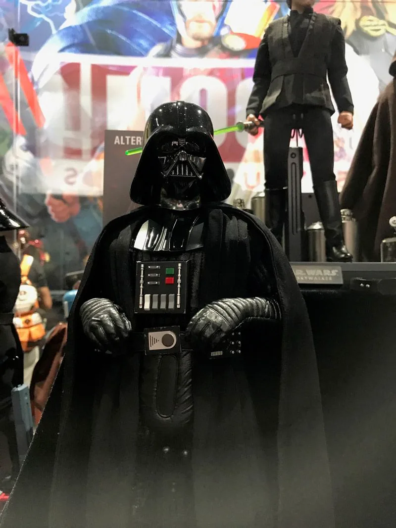 Hot Toys Darth Vader facing forward in black armor and cape, with both gloved hands raised near his illuminated-looking chest controls.