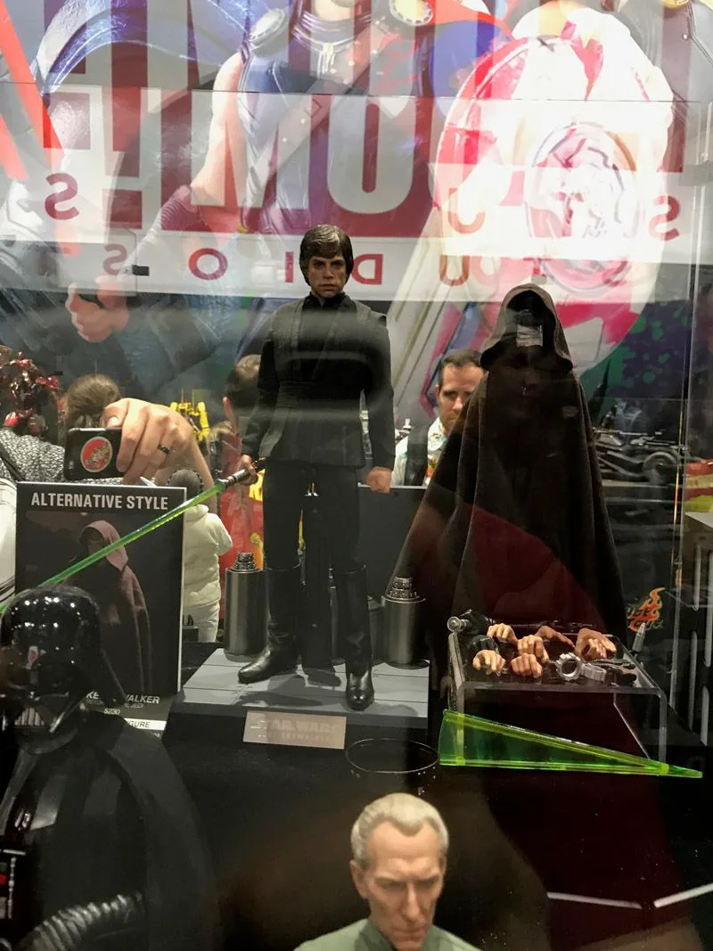 Hot Toys Luke Skywalker displayed in a black Jedi outfit beside a hooded presentation, with a green lightsaber and alternate hands nearby.