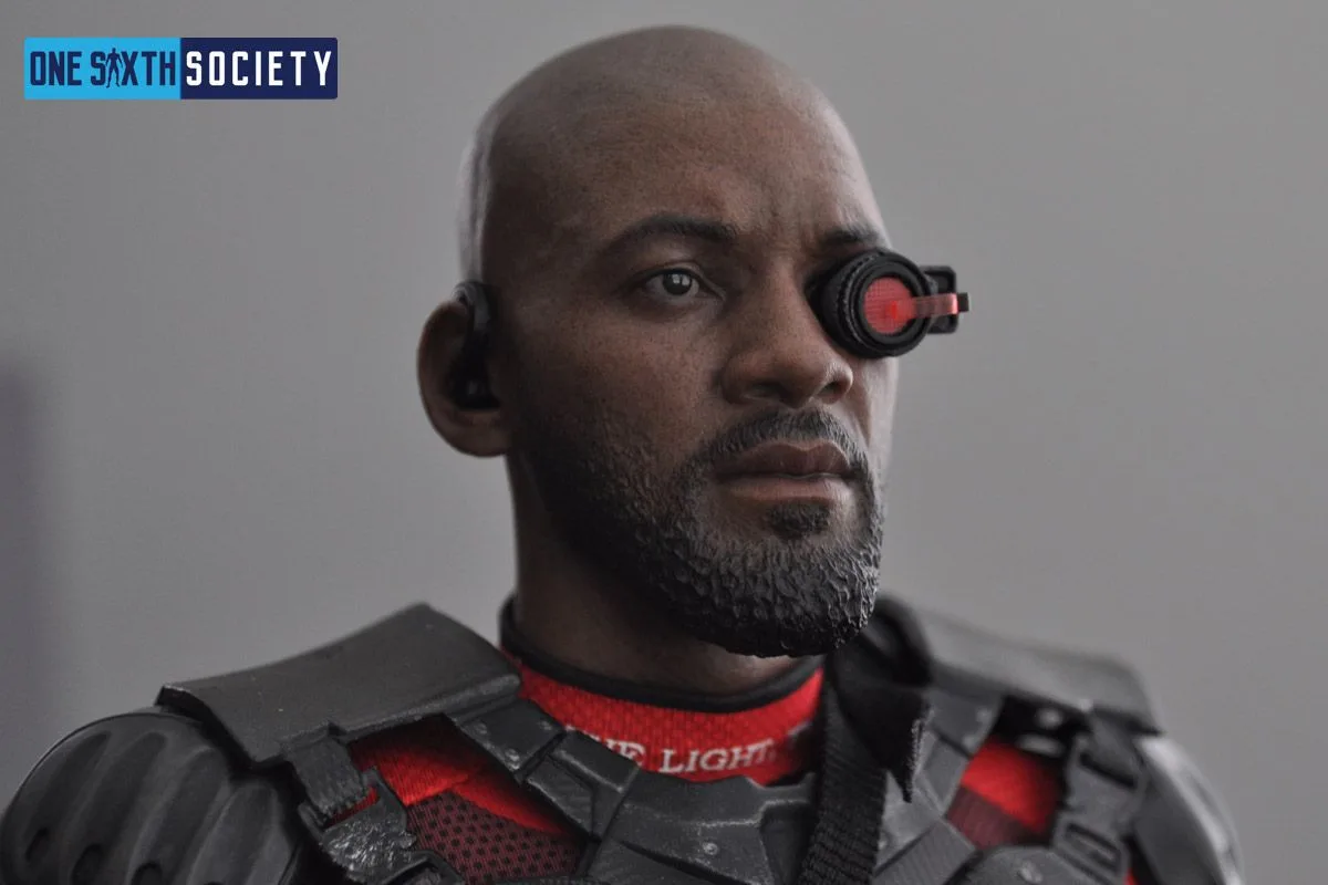 Close-up of the Hot Toys Deadshot unmasked head sculpt with painted beard, earpiece and red targeting scope over one eye.
