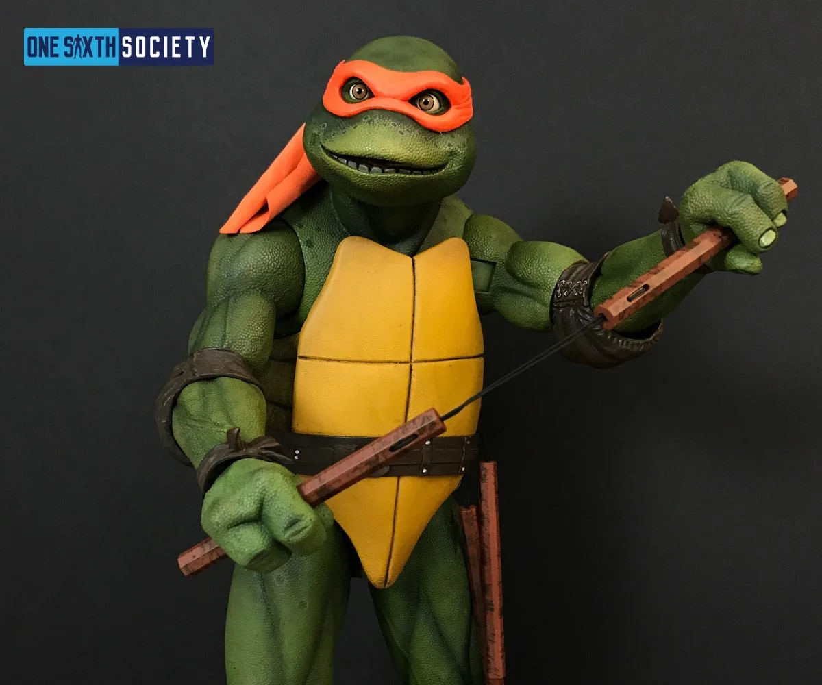 NECA Michelangelo holding one nunchuck handle raised and the other near his waist, with the connecting cord stretched across his torso.
