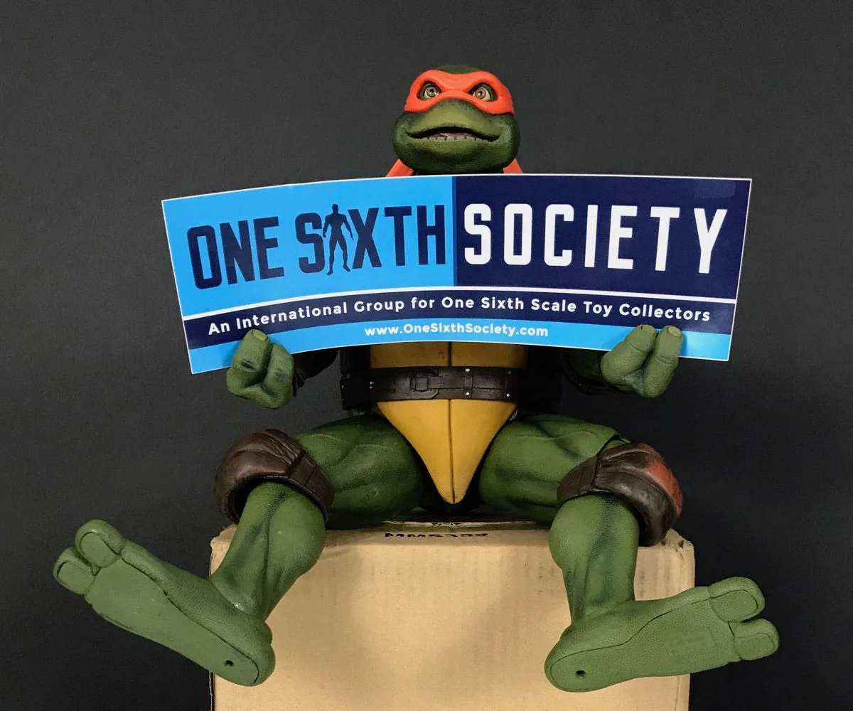 NECA Michelangelo seated on a cardboard box with his legs extended, holding a blue-and-black One Sixth Society sign.