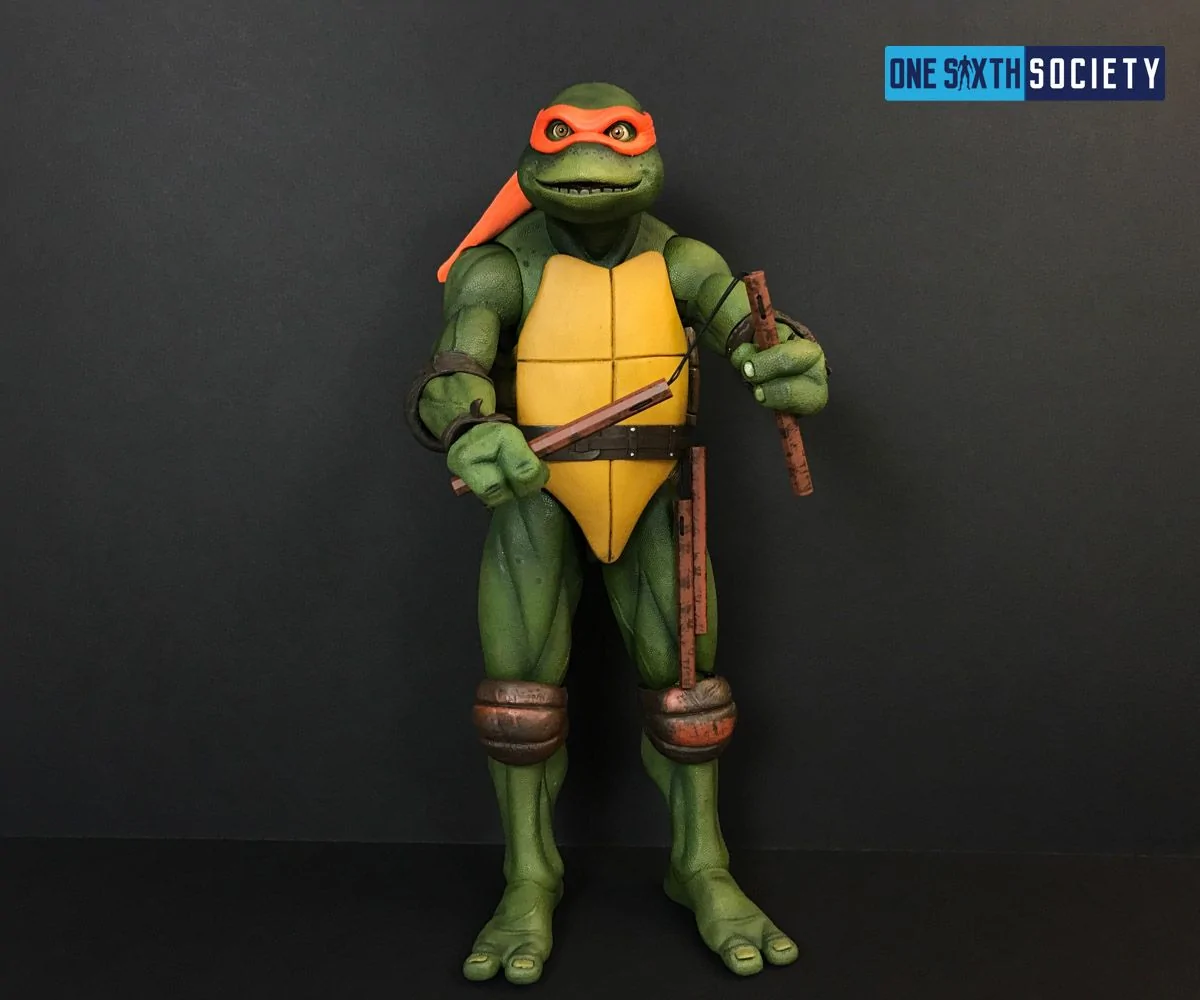 Full-length NECA movie Michelangelo figure in an orange mask holding wooden-look nunchucks against a dark backdrop.