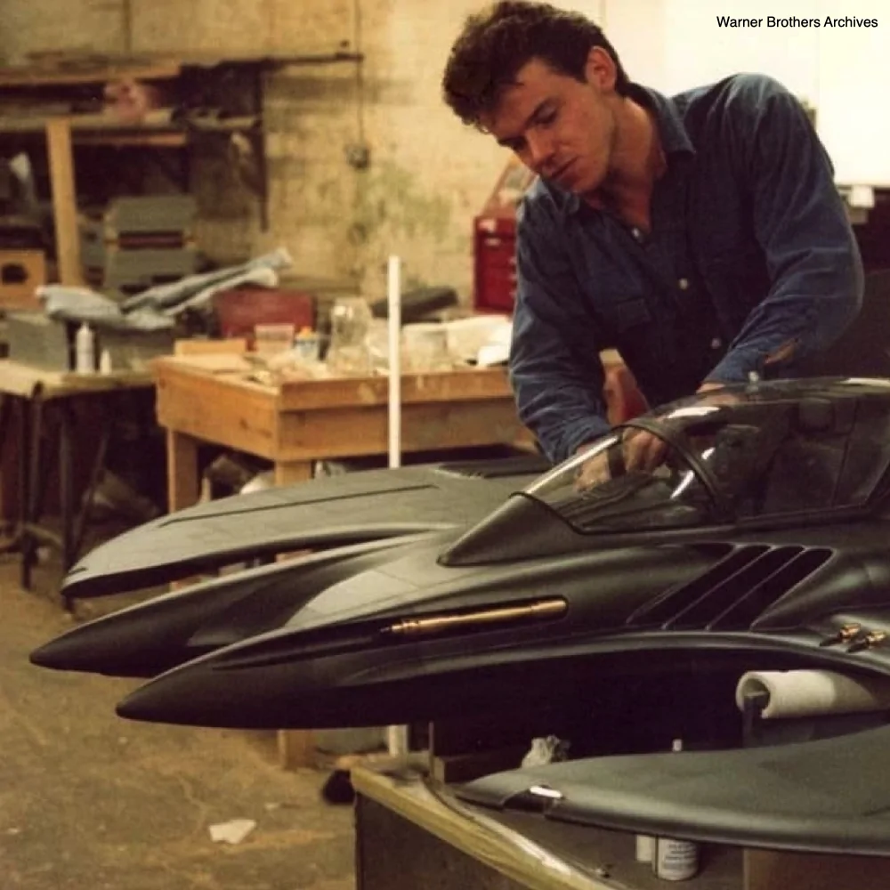 Archival workshop photograph credited to Warner Brothers Archives, showing a person working behind a large Batwing model on a bench.