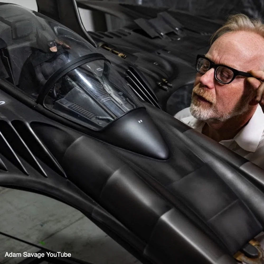 Image credited to Adam Savage’s YouTube channel, showing him looking closely at a large Batwing cockpit containing a Batman figure.