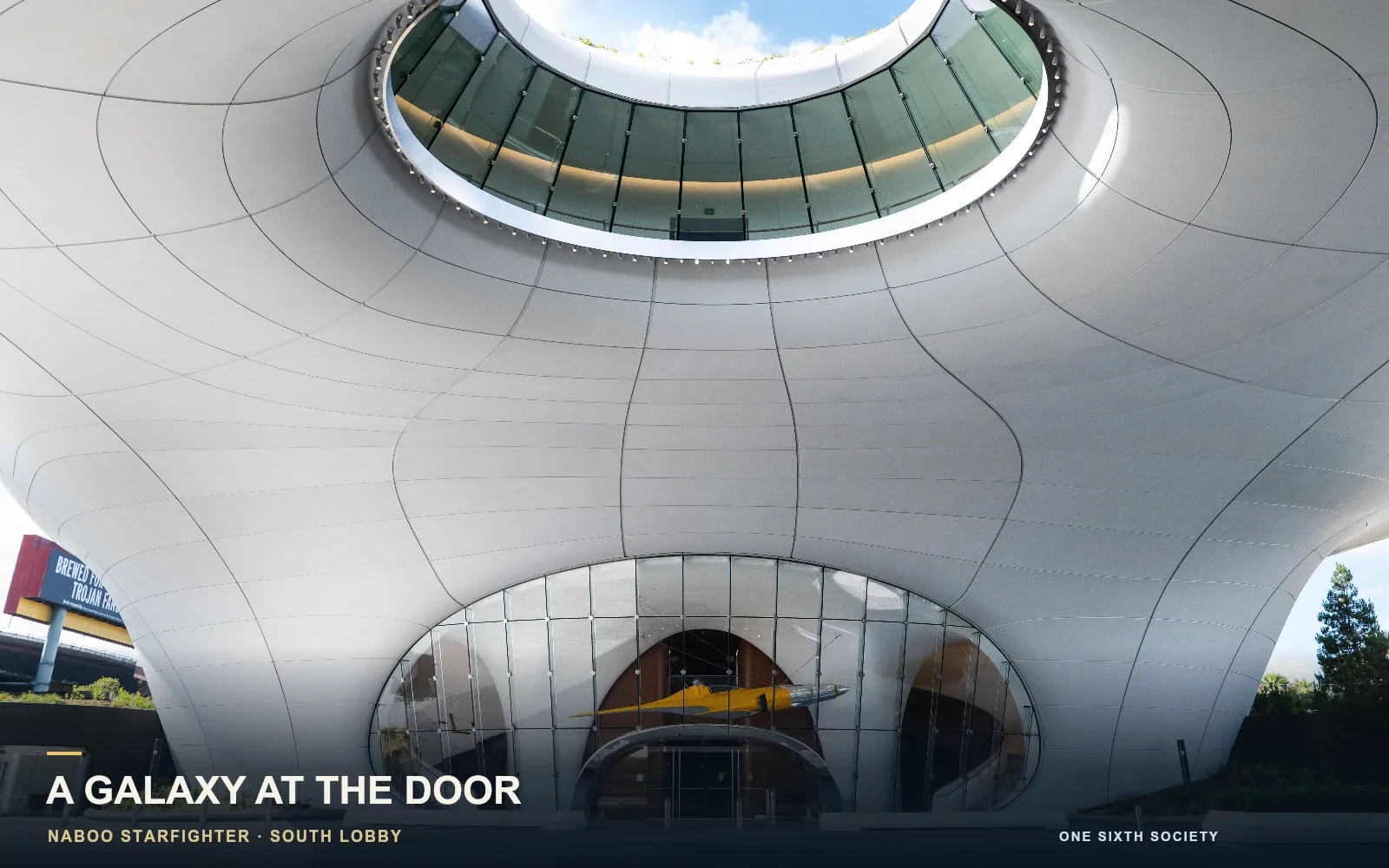 Yellow Naboo Starfighter visible through the Lucas Museum’s South Lobby glass entrance beneath a curved white overhang and circular skylight.
